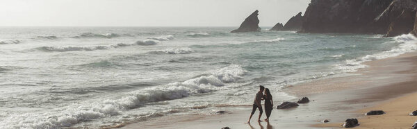 bearded man hugging girlfriend in dress while walking in ocean water, banner