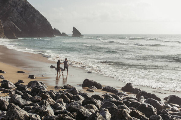 bearded man holding hands with tattooed girlfriend in dress while walking in ocean water 