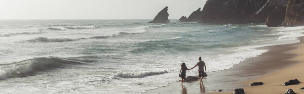 back view of man holding hands with girlfriend in dress while walking on wet sand near ocean water, banner