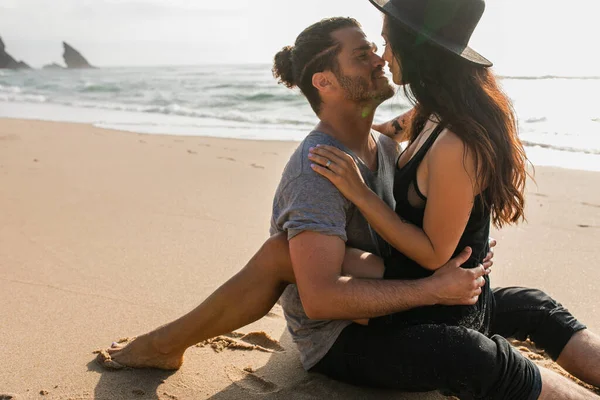戴着帽子的快乐而文身的女人与男朋友坐在沙滩上 — 图库照片