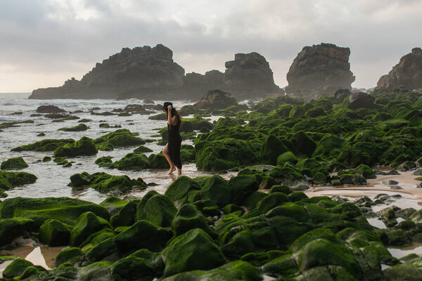 scenic view of woman in black dress and hat walking near mossy green stones in ocean