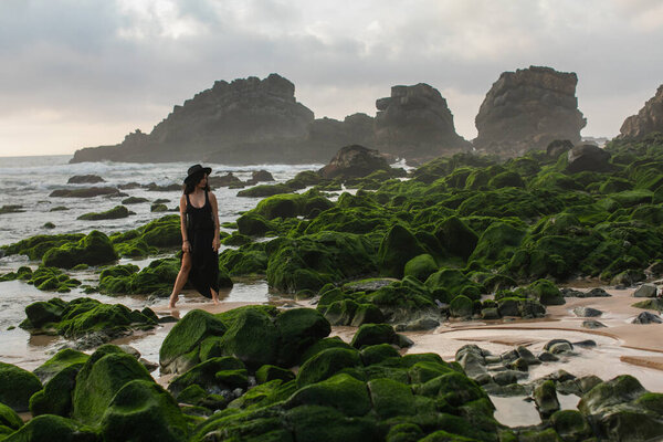 full length of tattooed woman in black dress and hat standing near mossy stones in ocean