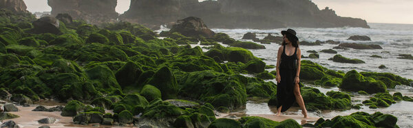 full length of tattooed woman in black dress and hat standing near mossy stones in ocean, banner