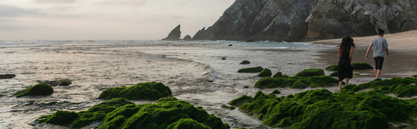 back view of couple walking near green mossy stones in ocean during sunset, banner