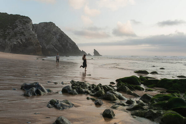 happy woman holding hat and running on wet sand near boyfriend  