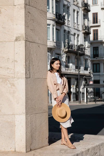 Vue pleine longueur de femme élégante avec chapeau de paille debout près du mur et regardant loin — Photo de stock