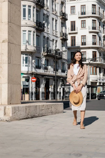 Full length view of brunette woman with straw hat walking on city street - foto de stock