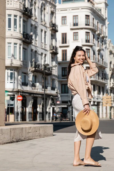 Full length of happy woman in trendy clothes holding straw hat on urban street — Stockfoto