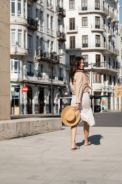 Mujer feliz en falda elegante caminando por la calle con sombrero de paja y sonriendo a la cámara - foto de stock