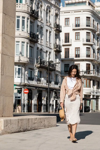 Full length of trendy woman with straw hat smiling while walking in city — стоковое фото