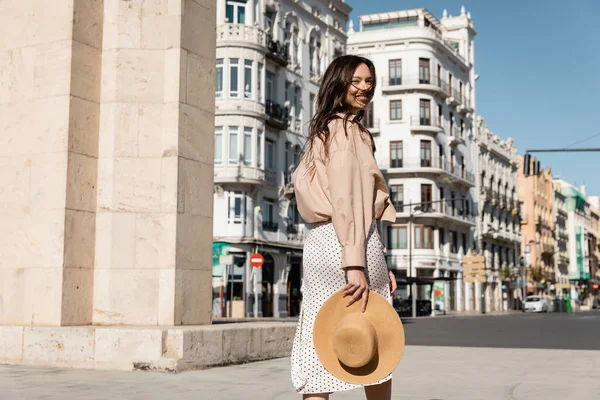 Cheerful woman with straw hat walking on windy street and looking at camera — Stock Photo