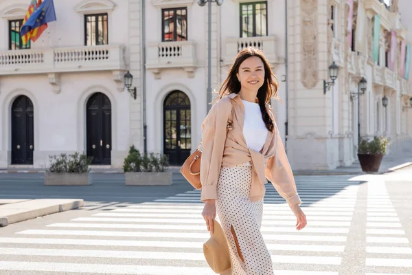 Stylish woman with handbag and straw hat smiling at camera near crosswalk — стоковое фото