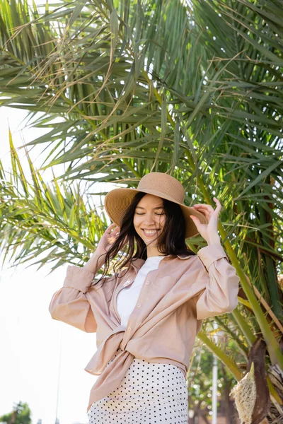 Cheerful brunette woman adjusting straw hat under palm tree — Stock Photo
