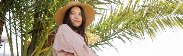 Smiling woman in straw hat looking away near green palm leaves, banner — стоковое фото