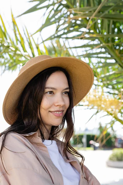 Portrait of brunette woman in straw hat smiling under blurred palm tree — Stock Photo