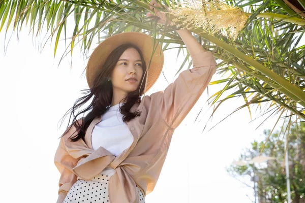 Visão de baixo ângulo da mulher morena na camisa bege e chapéu de palha sob ramos de palma verde — Fotografia de Stock