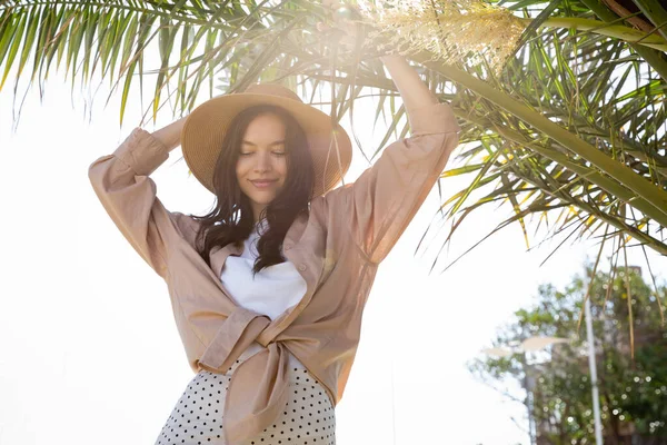Donna alla moda in camicia beige e cappello di paglia sorridente sotto foglie di palma verde — Foto stock