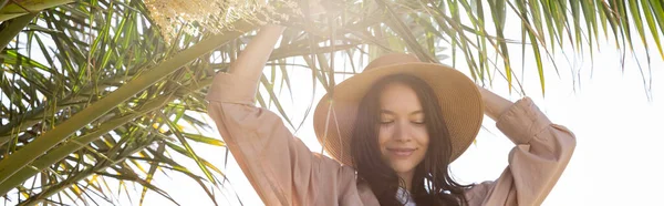 Pleased brunette woman in straw hat standing near green palm tree, banner — Stock Photo