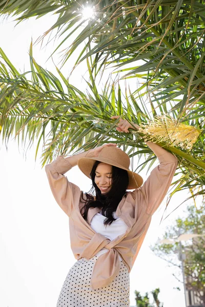 Joyful brunette woman in shirt and straw hat standing under green palm branches — Stock Photo