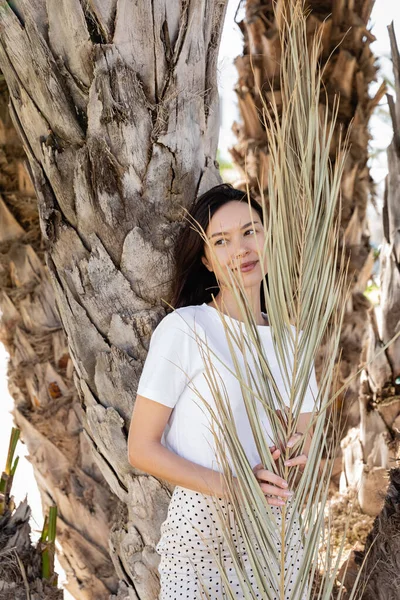 Felice donna bruna in t-shirt bianca in piedi vicino a tronchi di palma — Foto stock