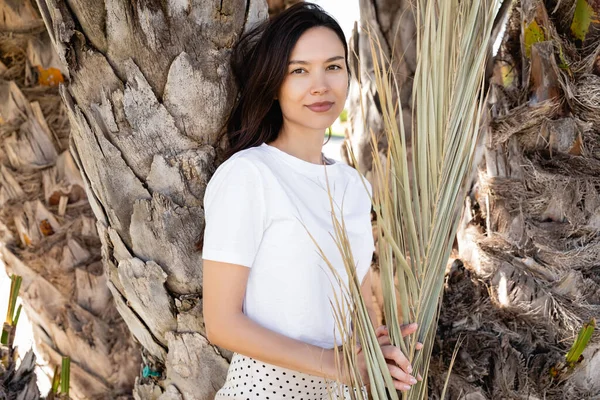 Brunette woman in white t-shirt smiling at camera near palm tree trunks — Stock Photo