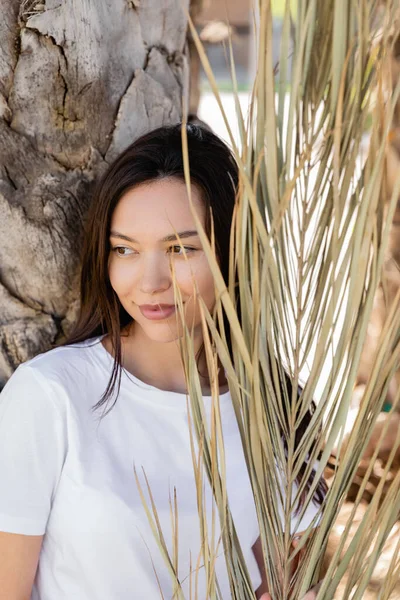 Brunette woman in white t-shirt smiling near dried palm leaves — Stock Photo