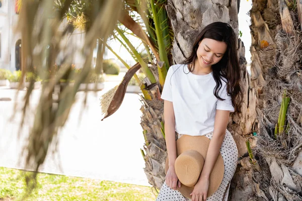 Smiling brunette woman holding straw hat while standing near palm tree — Stock Photo