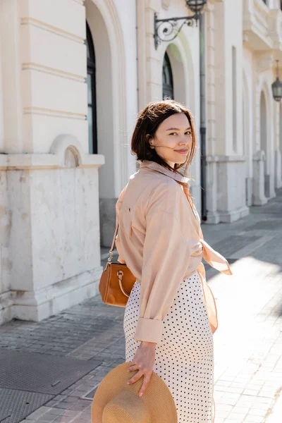 Donna bruna con borsetta e cappello di paglia sorridente alla macchina fotografica sulla strada della città — Foto stock