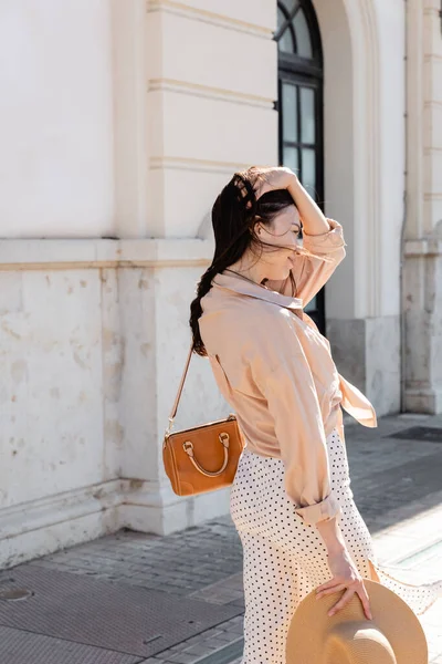 Brunette woman with handbag fixing hair while standing on street — Stock Photo