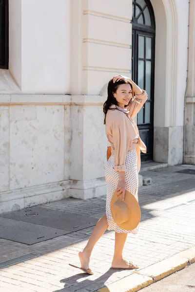 Happy woman fixing hair and looking at camera while walking on street with straw hat — Photo de stock