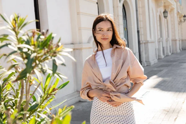 Positive brunette woman in trendy clothes standing near blurred plant on street — Stockfoto