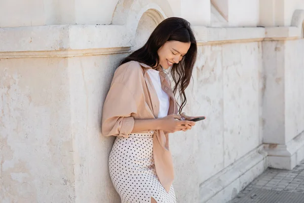 Mulher morena na moda sorrindo enquanto mensagens no smartphone perto da parede branca — Fotografia de Stock