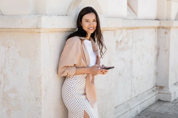 Mulher feliz e elegante de pé com smartphone perto da parede branca e olhando para a câmera — Fotografia de Stock