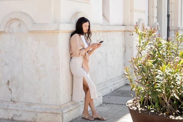 Comprimento total da mulher em mensagens saia no telefone móvel perto de edifício branco e planta verde — Fotografia de Stock