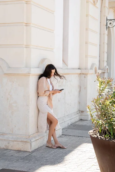 Full length view of stylish woman standing near white building and using smartphone — Stockfoto