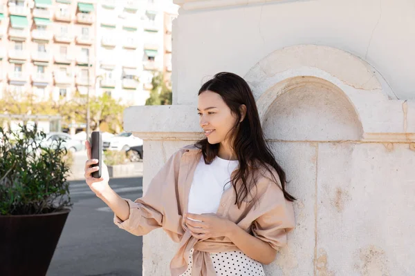 Smiling brunette woman taking selfie on smartphone near white wall on street — Stockfoto