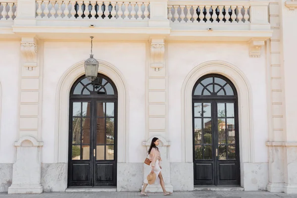 Side view of woman with handbag and straw hat walking along white building with arch doors — Photo de stock