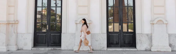 Side view of trendy woman walking along white building with black doors, banner — стоковое фото