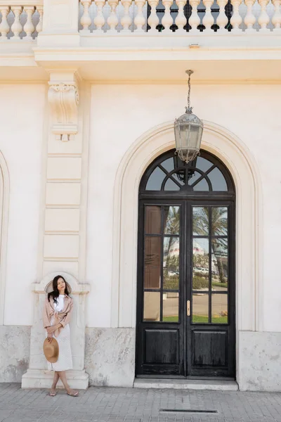 Longitud completa de la mujer morena de moda de pie cerca de edificio blanco con puerta de arco - foto de stock