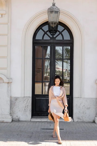 Full length of stylish brunette woman with handbag and straw hat walking on urban street — стоковое фото