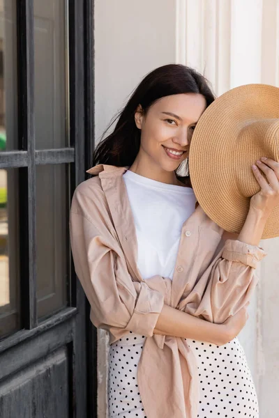 Woman in beige shirt obscuring face with straw hat and smiling at camera — Stockfoto