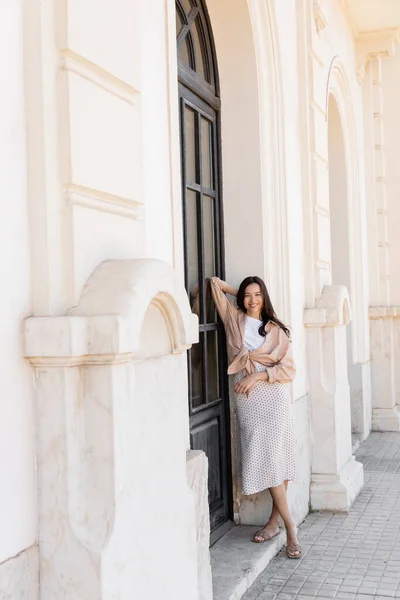 Comprimento total da mulher feliz de pé com a mão atrás da cabeça perto do edifício branco — Fotografia de Stock