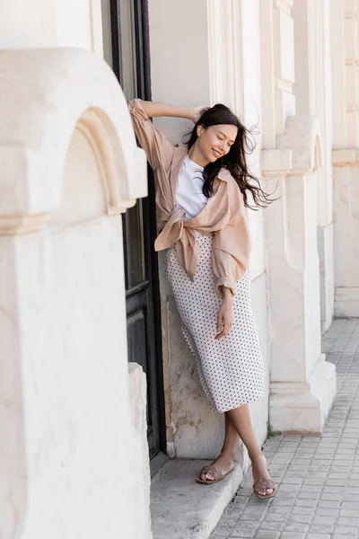 Full length view of happy woman in stylish skirt standing near white building — стоковое фото