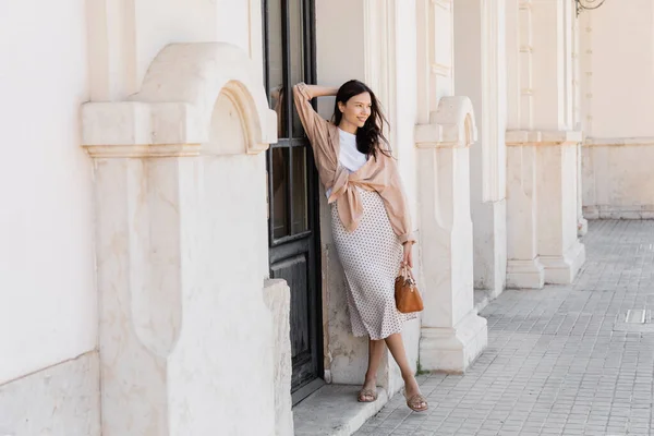 Повний вигляд усміхненої жінки з сумочкою, яка дивиться далеко, стоячи біля білої будівлі — стокове фото