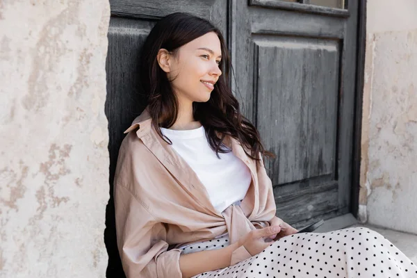 Smiling woman sitting with cellphone near grunge wooden door and looking away - foto de stock