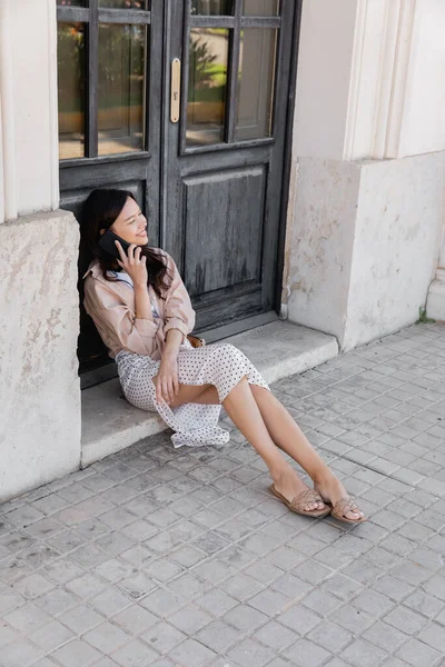 Visão de comprimento total da mulher feliz sentada perto de portas de madeira preta e falando no smartphone — Fotografia de Stock