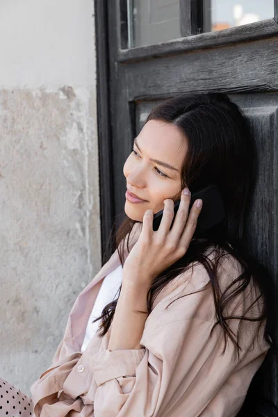 Улыбающаяся женщина в бежевой рубашке разговаривает по мобильному телефону на улице — стоковое фото