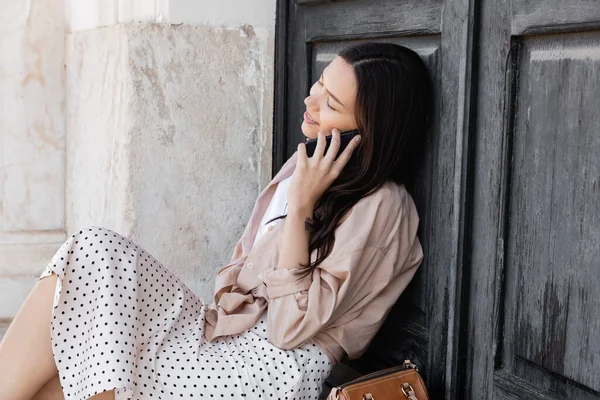 Lächelnde Frau mit geschlossenen Augen sitzt neben schwarzer Holztür und spricht auf Smartphone — Stockfoto