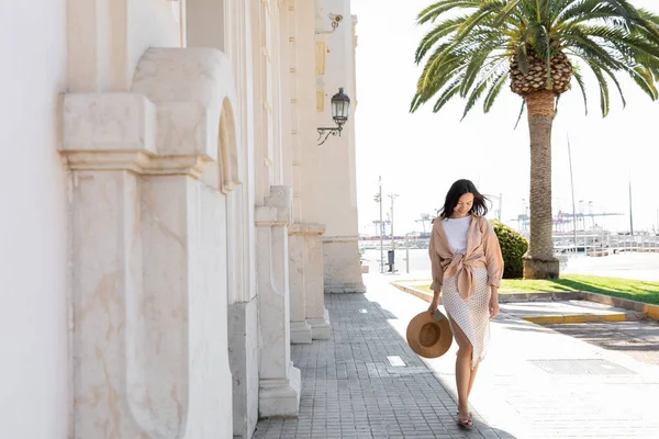 Full length of stylish woman walking on street with white building and palm tree — Stock Photo