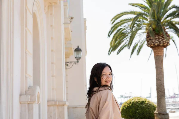 Brunette woman smiling at camera near white building and blurred palm tree — Stock Photo